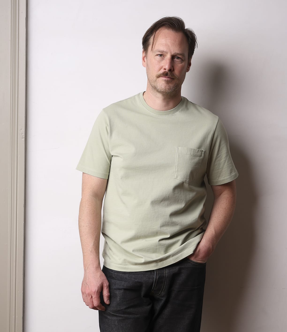 A man with light brown hair and a mustache stands against a plain wall, wearing the Best Made Co. Standard Pocket T-Shirt in Haze—a pale green garment-dyed tee with a chest pocket—and dark jeans, one hand in his pocket and a neutral expression.