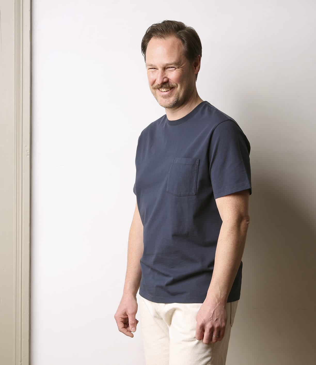 A man with light brown hair and a mustache smiles while wearing the Best Made Co Standard Pocket T-Shirt in navy and light-colored pants, standing against a plain light wall.