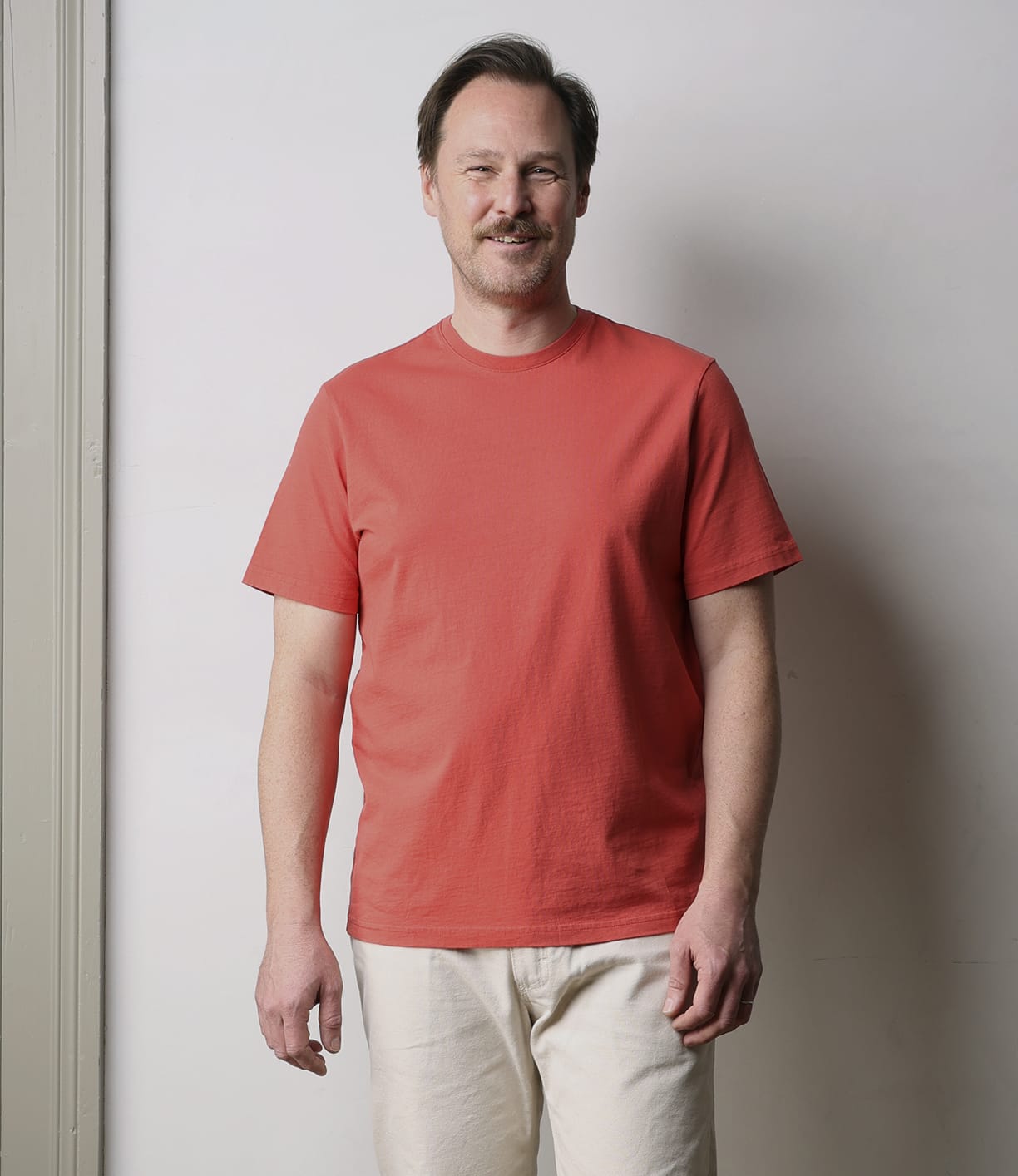 A man with short brown hair and a mustache stands smiling against a plain light wall, wearing the Best Made Co. Standard T-Shirt in faded red—an everyday essential, paired with light beige pants.