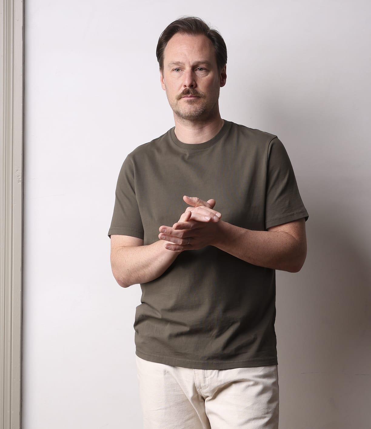 A man with light brown hair and a mustache wears the Best Made Co. Standard T-Shirt Field Drab, made from Peruvian tangüis cotton, and light beige pants, standing against a plain white wall with his hands clasped, looking forward.
