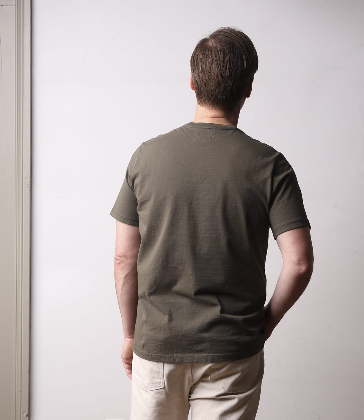A person with short brown hair, wearing the Best Made Co. Standard T-Shirt in Field Drab and light pants, stands facing away from the camera against a plain light-colored wall.