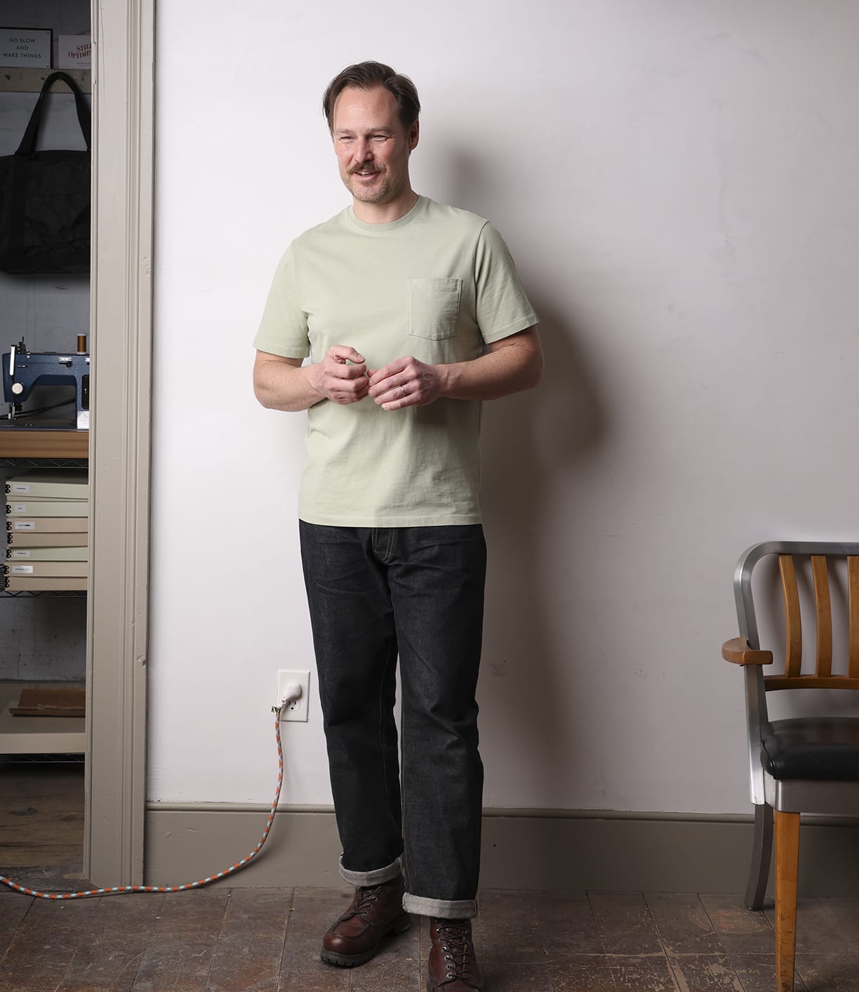 A man wearing the Best Made Co. Standard Pocket T-Shirt Haze stands indoors near a wooden chair, smiling with his hands together. A sewing machine and a coiled extension cord are visible in the background.