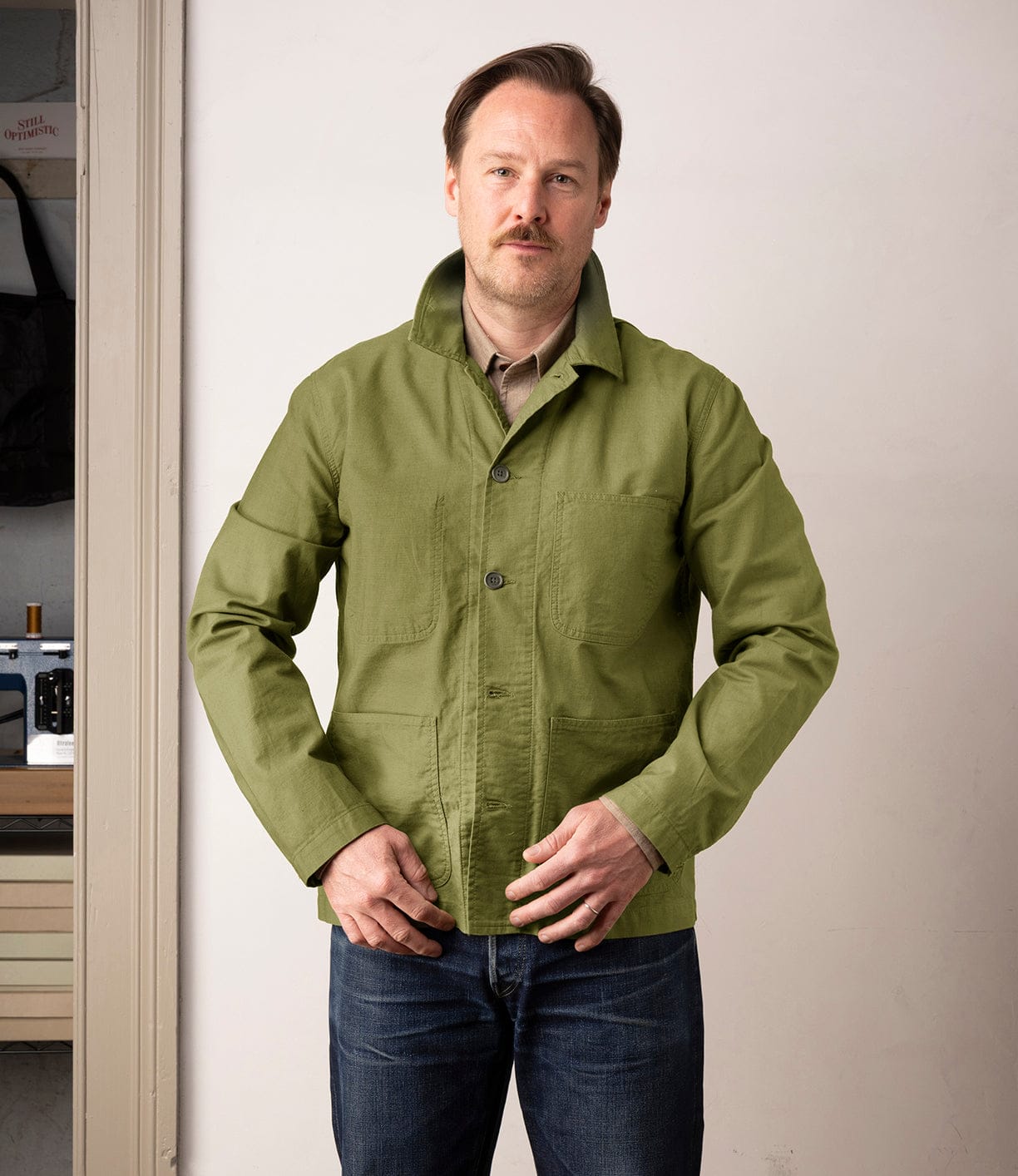 A man with light brown hair and a mustache stands indoors, wearing the Best Made Co. Panama Cloth Service Jacket over a tan shirt and blue jeans, looking at the camera while adjusting his jacket.