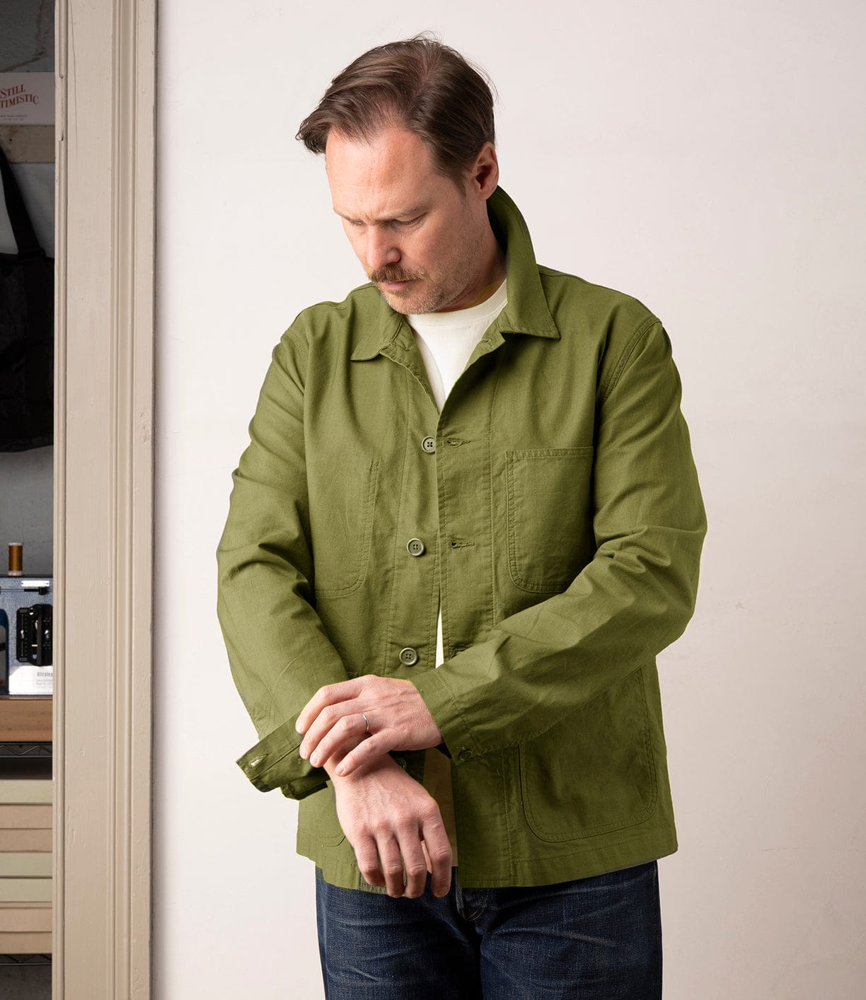 A man with light brown hair and a short beard fastens the cuff of his olive green Panama Cloth Service Jacket by Best Made Co., made from Japanese cotton Panama cloth, worn over a white shirt with blue jeans while standing indoors near a doorway.