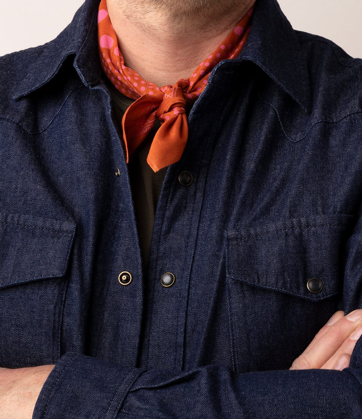 A person wearing the Best Made Co Fukuyama Snap-Front Shirt with a red-orange scarf with pink polka dots tied around their neck, arms crossed and face partially out of frame.