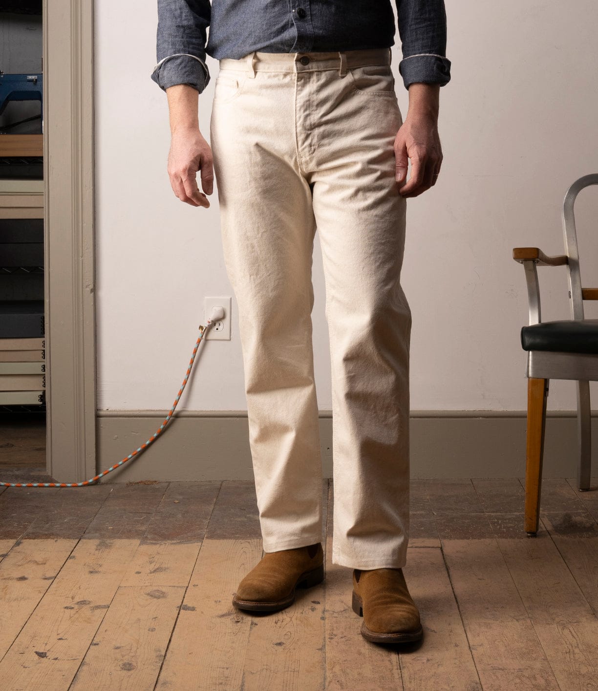 A person in a blue shirt, Best Made Co Standard 5-Pocket Pant M2 Undyed, and brown boots stands indoors on a wooden floor near a gray chair and an outlet with an orange and white cord plugged in.