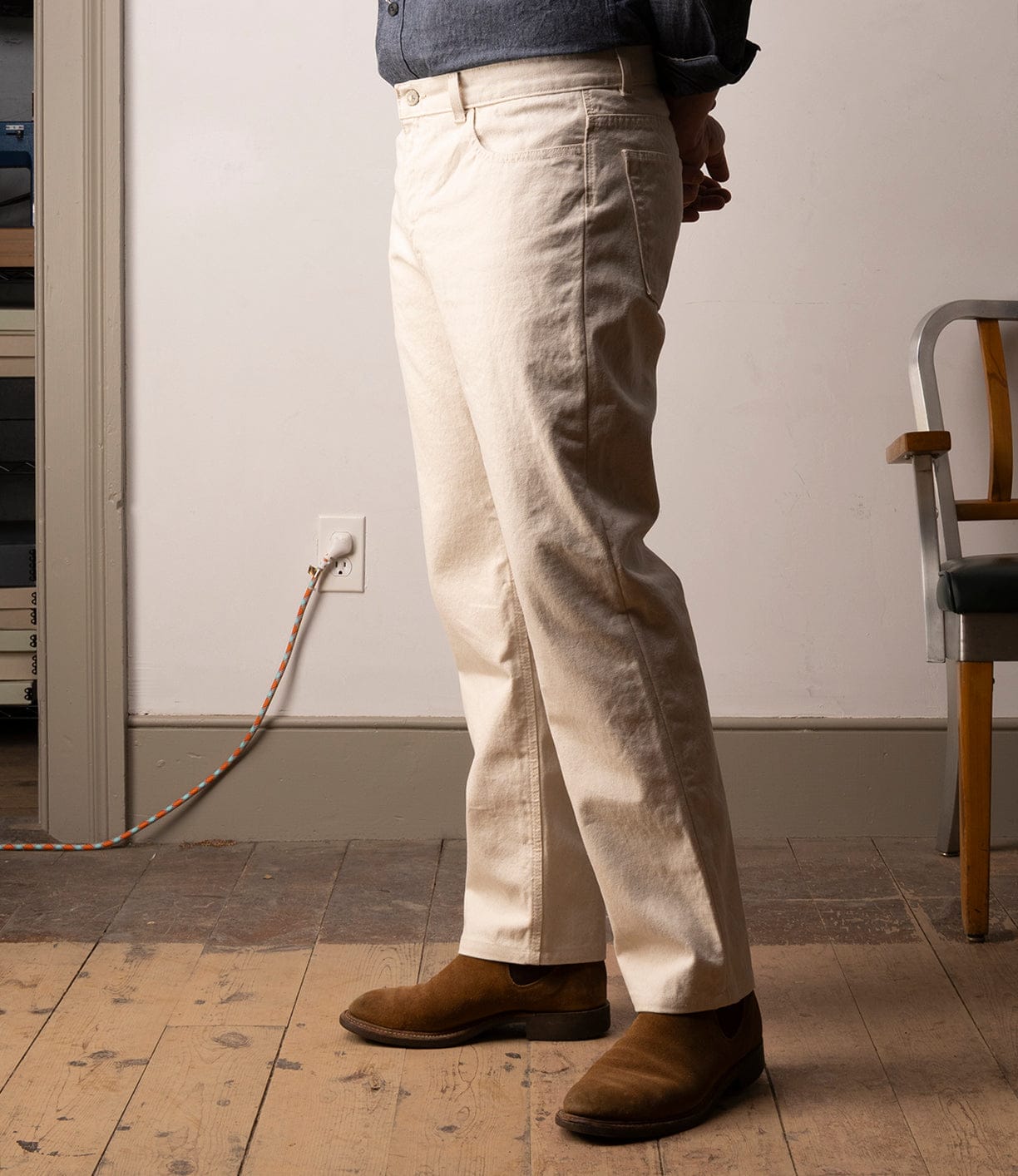 A person wearing Best Made Co Standard 5-Pocket Pant M2 Undyed, a dark shirt, and brown suede shoes stands indoors on wood flooring near a chair and wall outlet with an orange-and-white cord. The head is not visible.