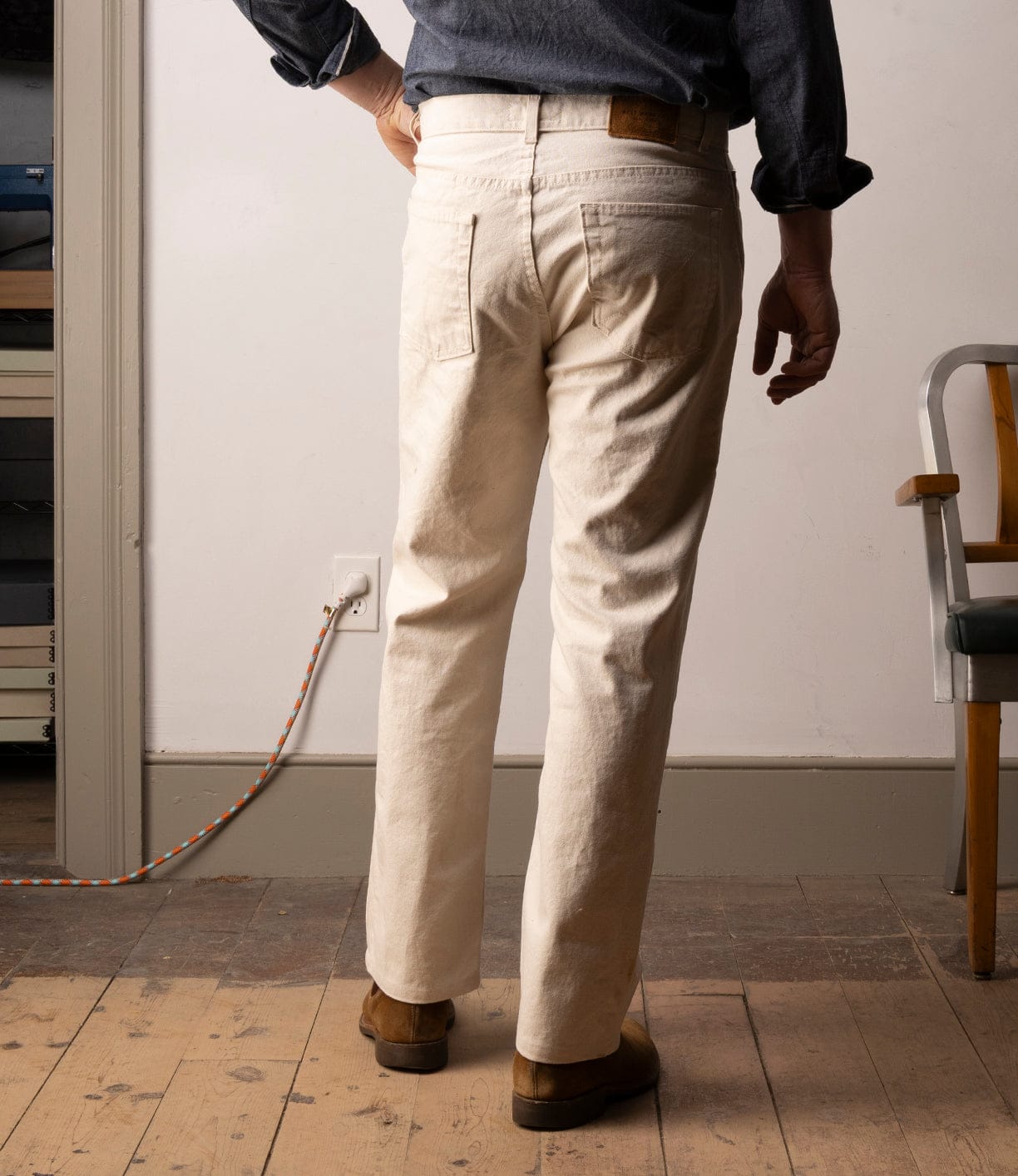 A person stands indoors facing away, wearing Best Made Co’s Standard 5-Pocket Pant M2 Undyed jeans and a denim shirt, paired with brown shoes. An orange-and-white extension cord is plugged into the wall; a wooden chair appears on the right.
