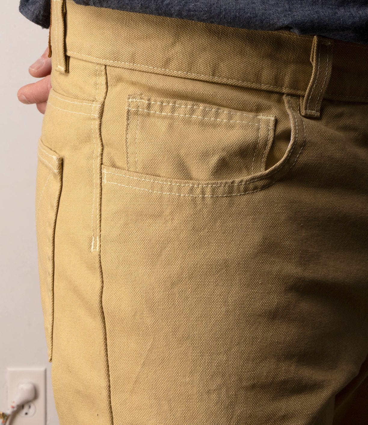 Close-up of a person wearing Best Made Co Standard 5-Pocket Pant M2 Khaki, showing front pocket, belt loop, white stitching, and part of their hand and gray shirt. A wall and electrical outlet are visible in the background.