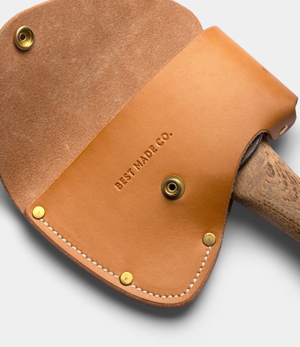 Close-up of a tan leather sheath with brass rivets and snaps, stamped with Best Made Co., partially covering the Appalachian hickory handle of the Spark (Straight-Hold Edition) campfire axe, on a light grey background.