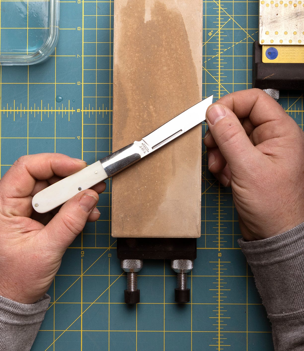 Someone sharpens a Best Made Co Barlow Knife, with a stainless steel blade, on a rectangular whetstone placed on a green cutting mat with yellow grid lines, holding the knife at an angle with both hands.