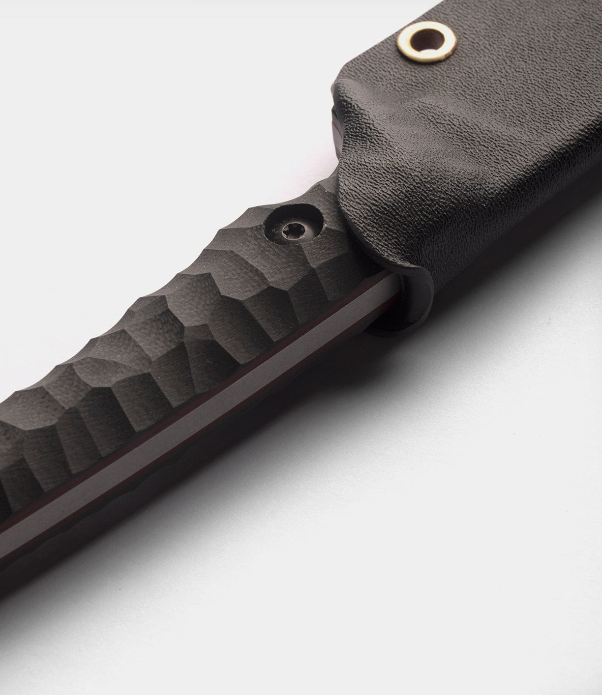 Close-up of the NWBW Field Knife (BMC Edition) by Best Made Co: black textured handle, partially covered by a black sheath with brass eyelet, on a plain light gray background. Made with durable American D2 tool steel.