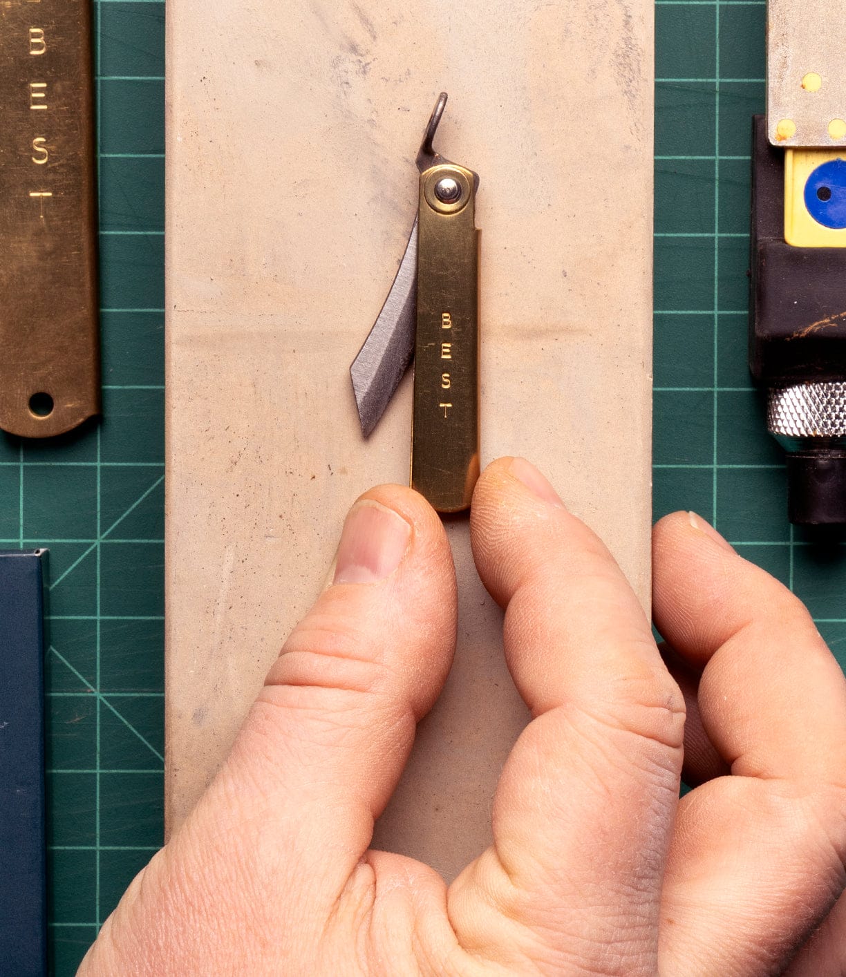 A hand holds a Mini Brass Higo Knife by Best Made Co on a stone sharpening block, with other tools partly visible on a green cutting mat.