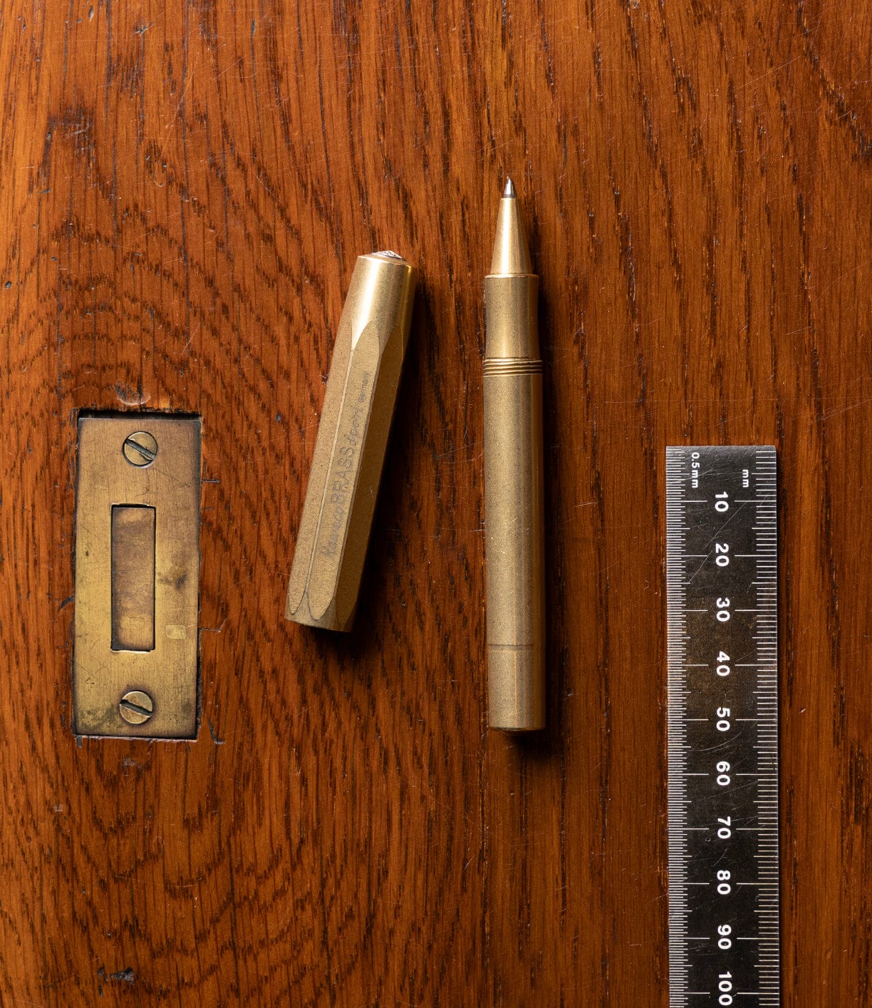 A Kaweco Sport brass pocket pen by Best Made Co., with its hexagonal cap off, rests beside a metal ruler showing precise centimeter and millimeter markings on a wooden surface. The pen is compatible with a G2 rollerball refill.