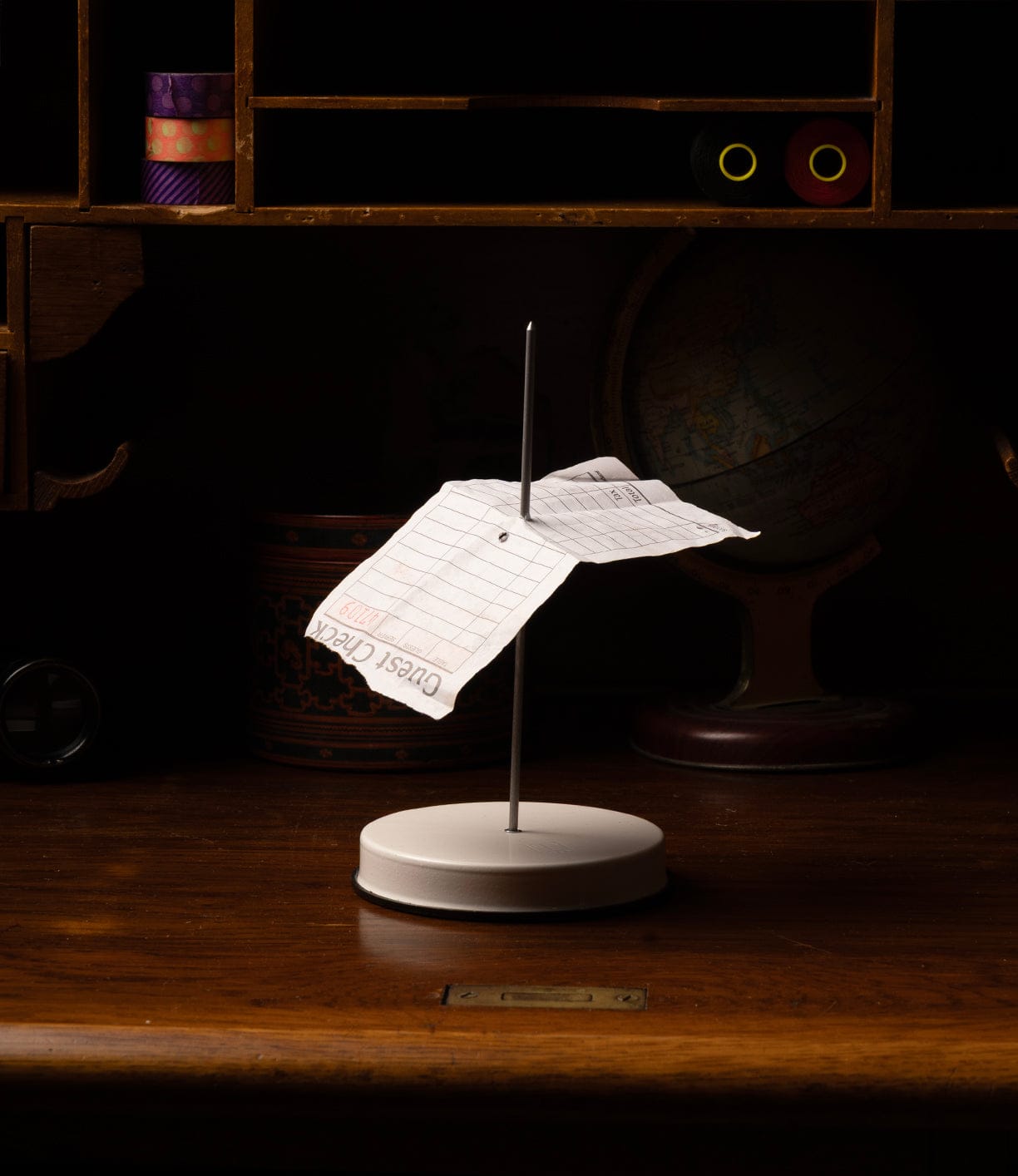 A paper receipt is impaled on the Best Made Co. Italian Receipt Spike, which sits on a wooden desk organizer with shelves, a globe, and other office items in the background.