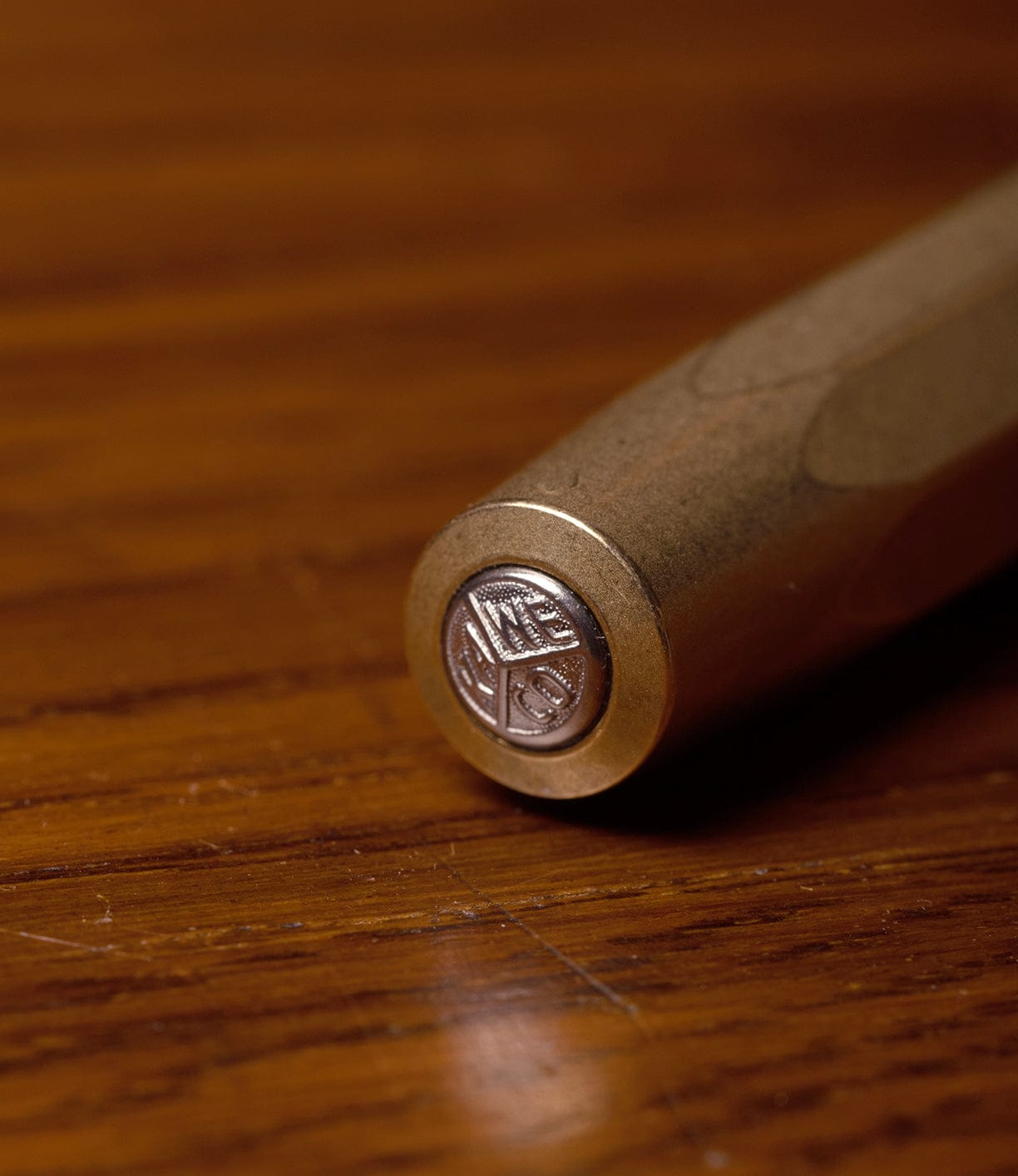 A close-up of the end of a Best Made Co. Kaweco Sport brass pocket pen rests on wood, displaying a circular emblem engraved with the letters IN E K S.