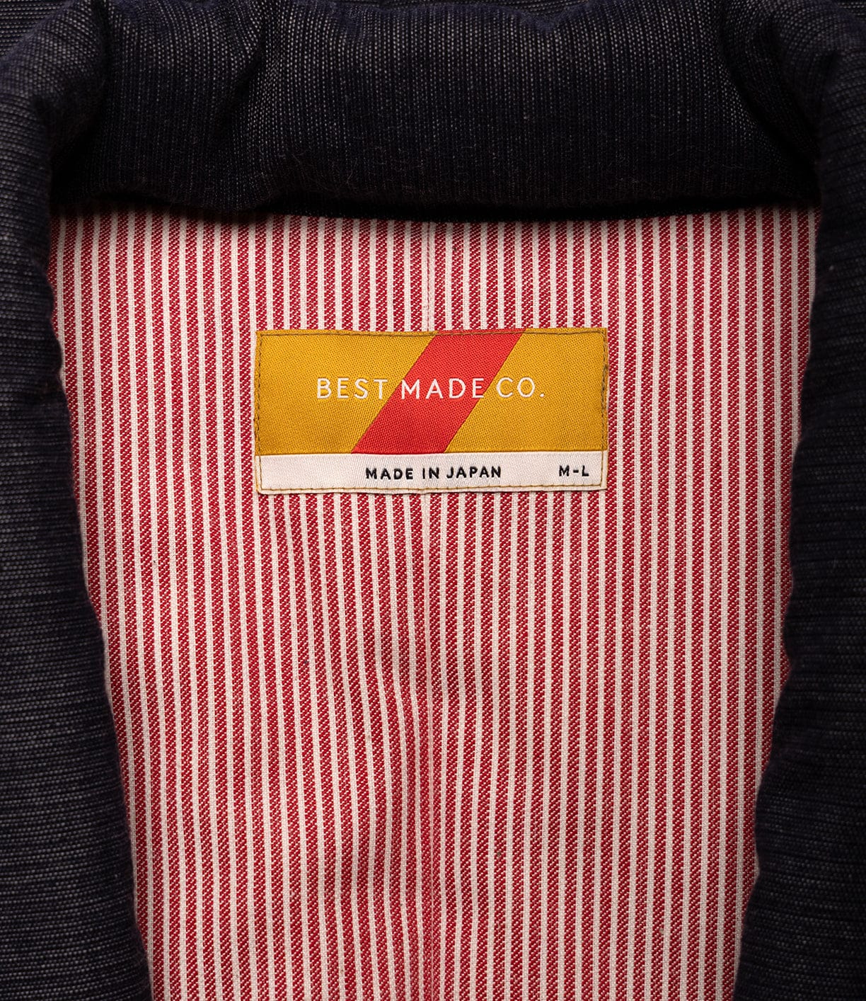 A close-up of the label inside a red and white striped Quilted Hanten Jacket by Best Made Co., made in Japan. The label, inspired by Japanese workwear, features a yellow and red diagonal design and displays the size M-L.