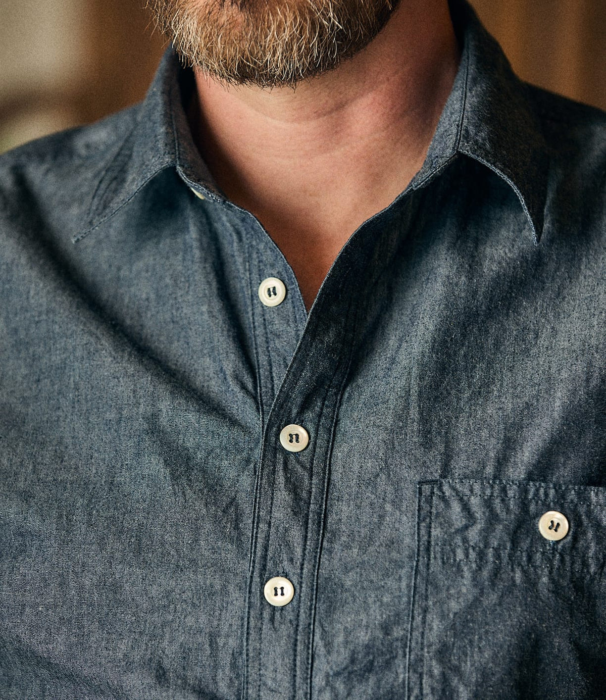 Close-up of a person wearing the Best Made Co. Indigo Chambray Popover, a blue button-up shirt made from premium Fukuyama woven cotton with white buttons and a front pocket. The wearer’s beard and part of their neck are visible.