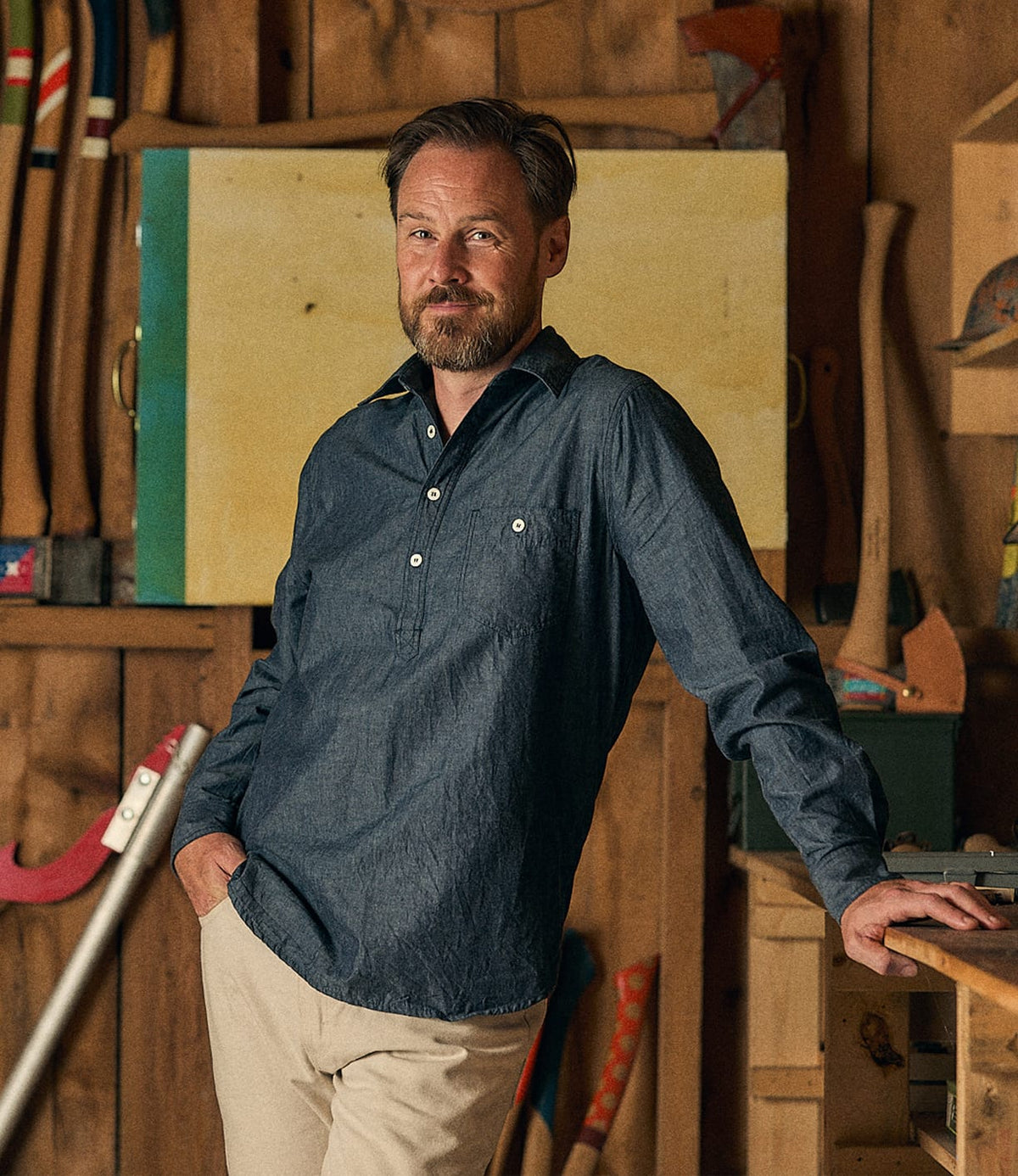 A bearded man wearing the Best Made Co. Indigo Chambray Popover and beige pants leans against a workbench in a wooden workshop, with axes and tools hanging on the wall behind him.