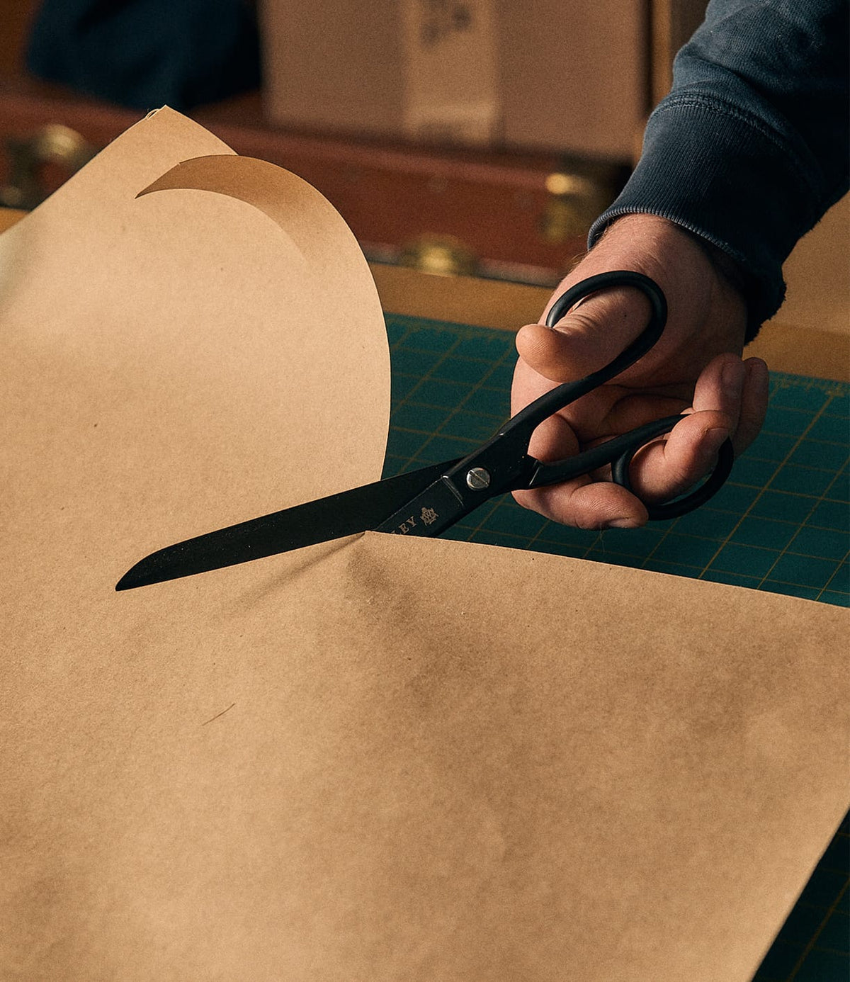 A person uses Best Made Co. Ten-Inch Paper Scissors to cut a large sheet of brown paper on a green cutting mat.