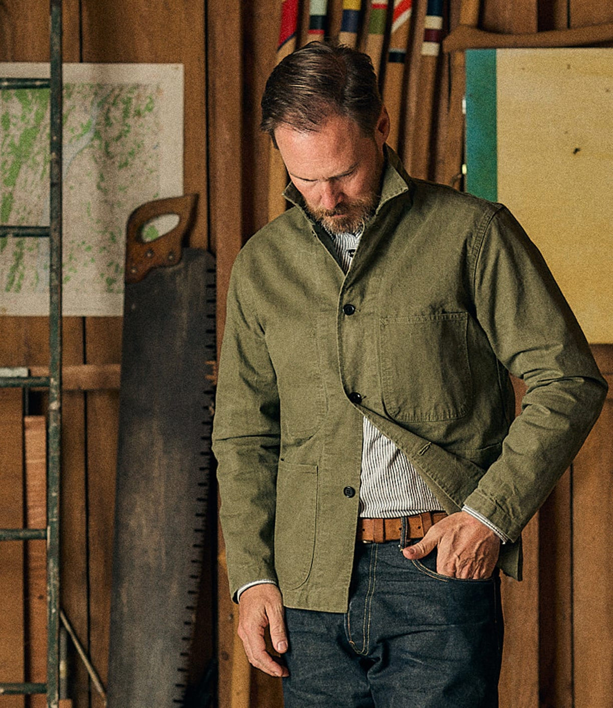 A man wearing the Best Made Co. Service Jacket Special Edition Olive Drab, crafted from Japanese twill, stands in a workshop with wooden walls, tools, and a ladder in the background—an homage to the utilitarian heritage of the classic P-41 Marine Corps style.