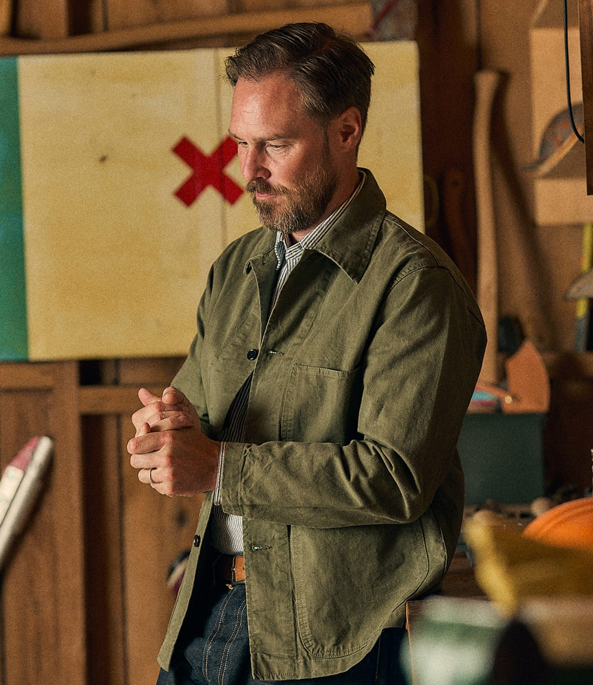 A bearded man stands indoors with his hands clasped, looking down, wearing the Best Made Co. Service Jacket Special Edition Olive Drab over a collared shirt. The background includes tools, a wooden wall, and a painting marked with a red X.