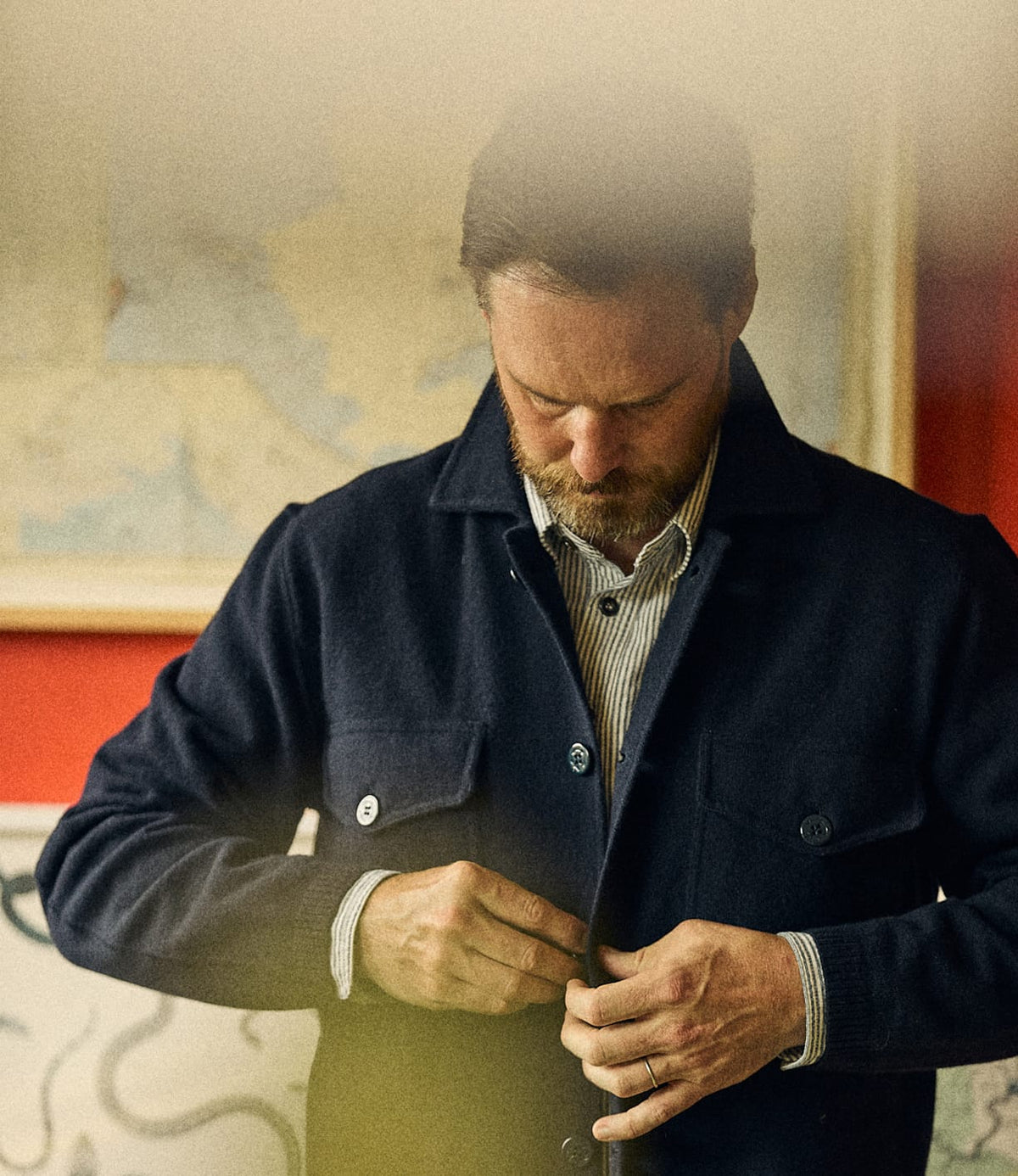 A bearded man buttons a navy Best Made Co. Knit Lambswool CPO shirt over a striped collared shirt.