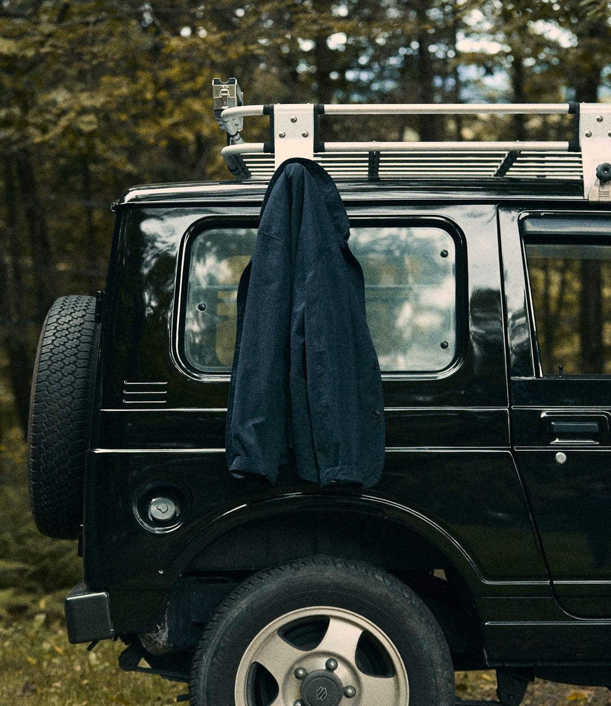 A Best Made Co. Possibilities Jacket Navy hangs on the roof rack of a black SUV parked in a forest, with trees in the background and a spare tire mounted on the back of the vehicle.