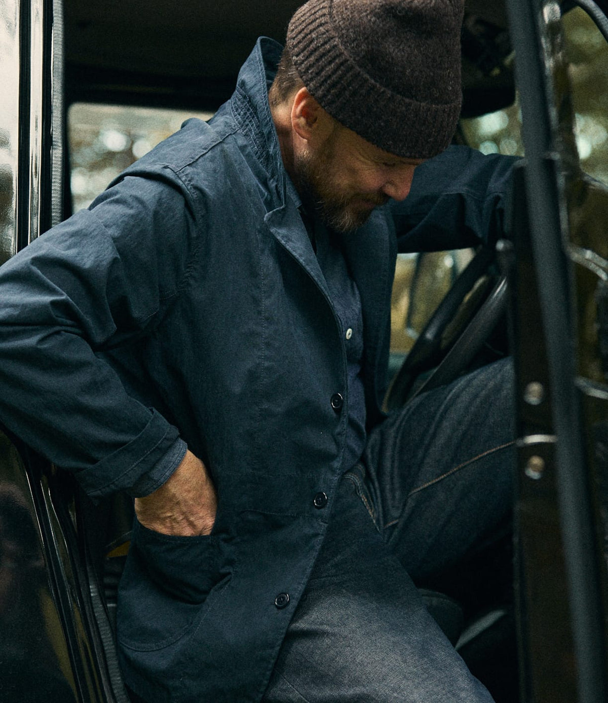 A person wearing a brown knit beanie and the Best Made Co. Possibilities Jacket Navy is stepping out of a vehicle, looking down and smiling slightly, with one hand in their pocket.
