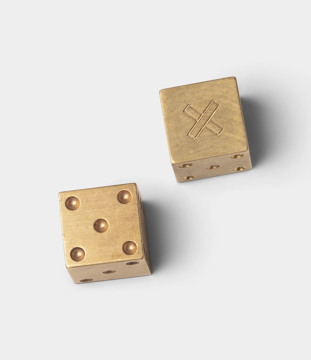 Two Best Made Co. Solid Brass Dice rest on a light gray surface—one shows four pips, while the other features a machined brass face with an engraved X in place of standard dots.