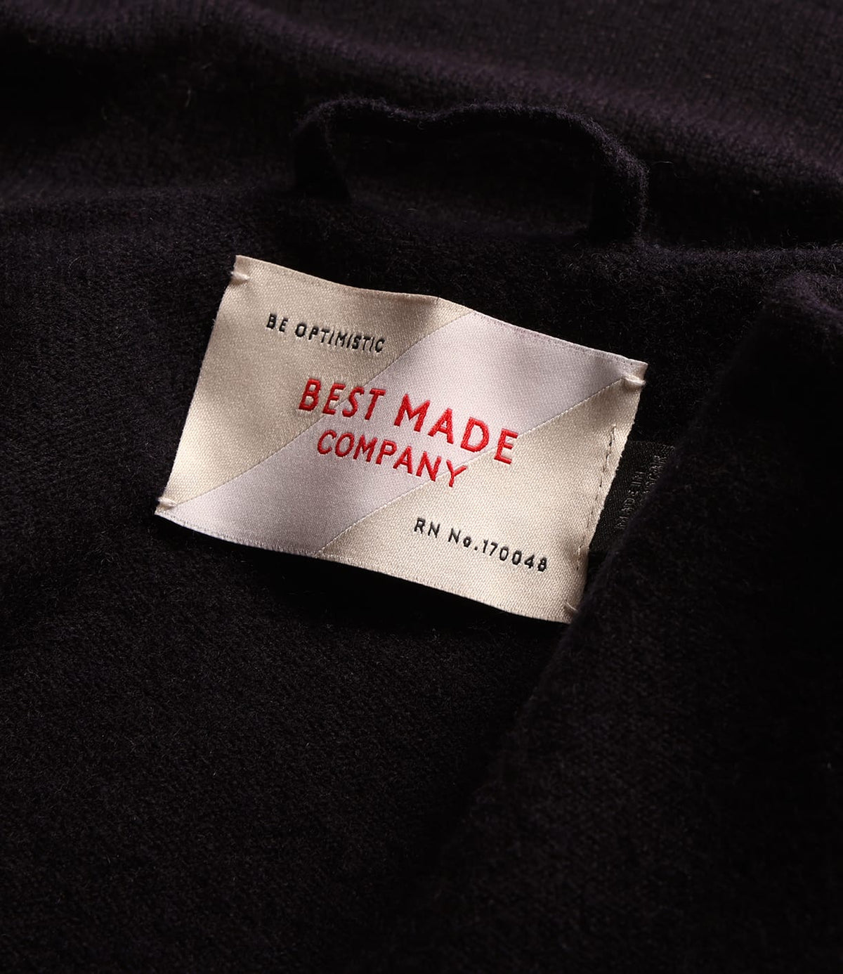 Close-up of a clothing label on a dark merino wool Knit Lambswool CPO shirt, reading Be Optimistic, Best Made Company, and RN No. 170048.