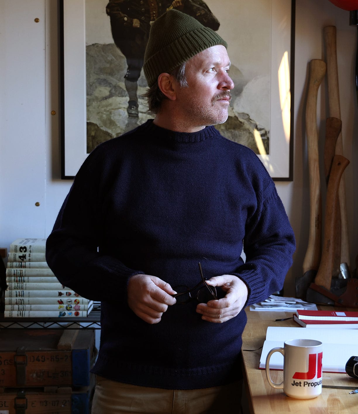 A man in a dark Best Made Co. Guernsey Sweater and green knit hat stands indoors by a desk with books, a mug, notebooks, and an axe, holding glasses and looking to the side. A framed artwork hangs on the wall behind him.