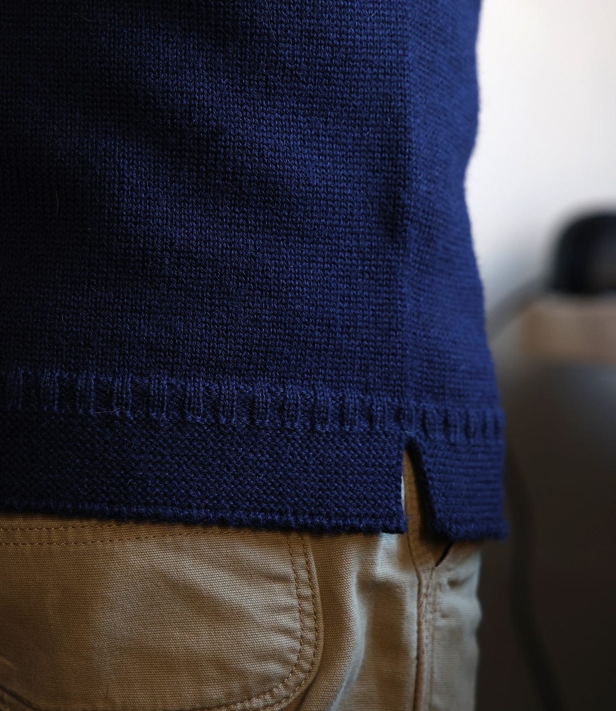 A close-up of a person wearing the Best Made Co. Guernsey Sweater in navy blue, made with traditional seamless knitting and a small side slit at the hem, paired with beige pants against a blurred background.