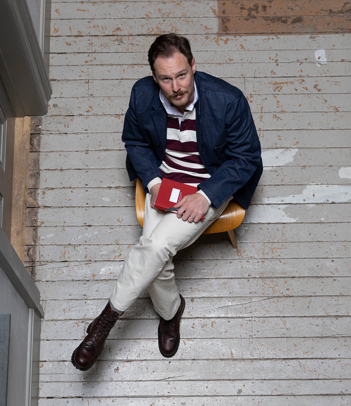 Man sitting on a wooden chair holding a red book on a wooden floor.