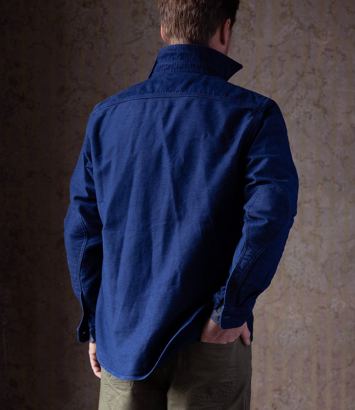 A person faces away from the camera, wearing the Best Made Co Indigo Moleskin Work Shirt with a raised collar and olive green pants, set against a textured, muted wall.