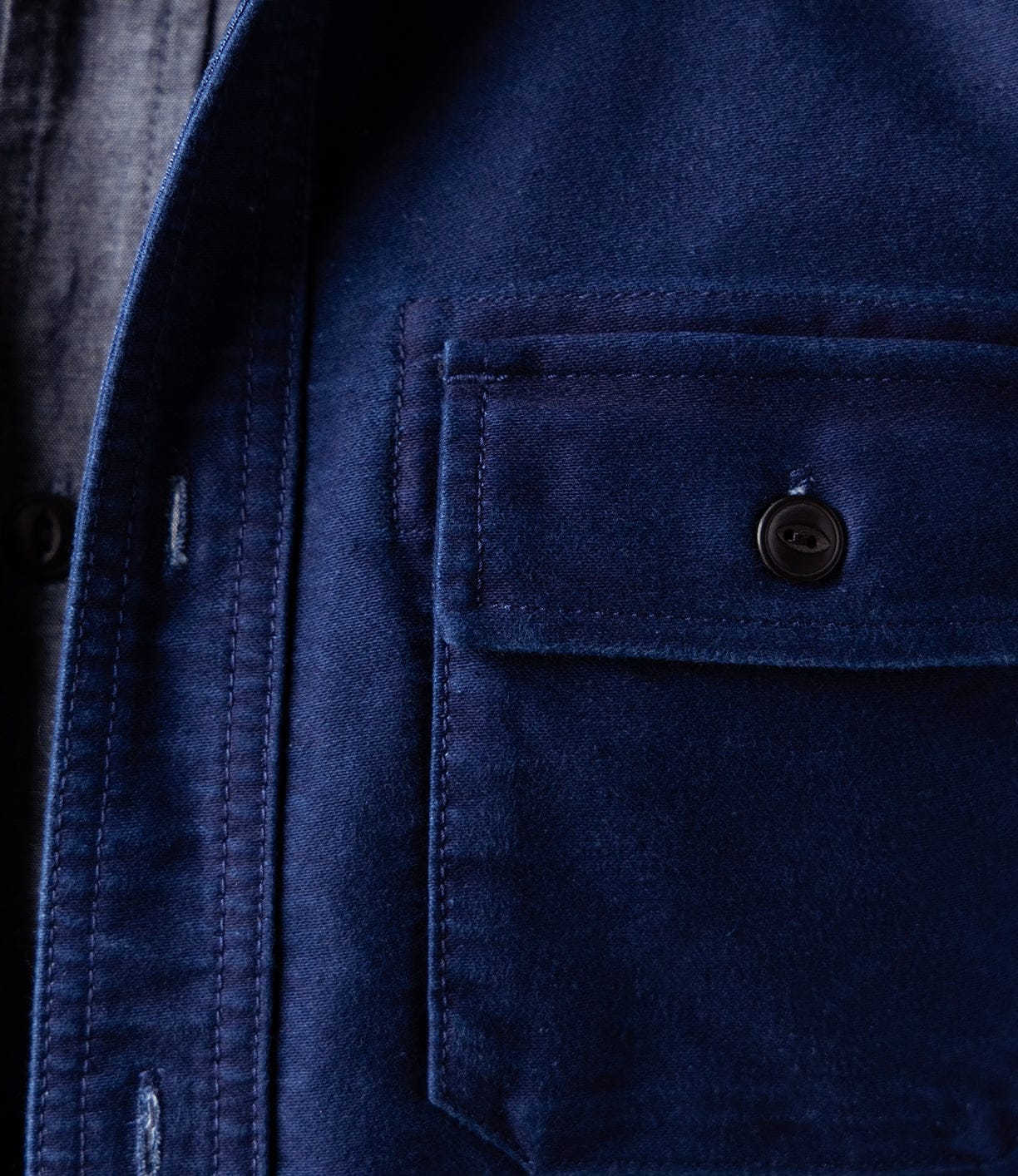 Close-up of the Best Made Co Indigo Moleskin Work Shirt, highlighting its buttoned chest pocket, intricate stitching, and rich texture. A hint of a grey shirt underneath appears on the left side of the image.