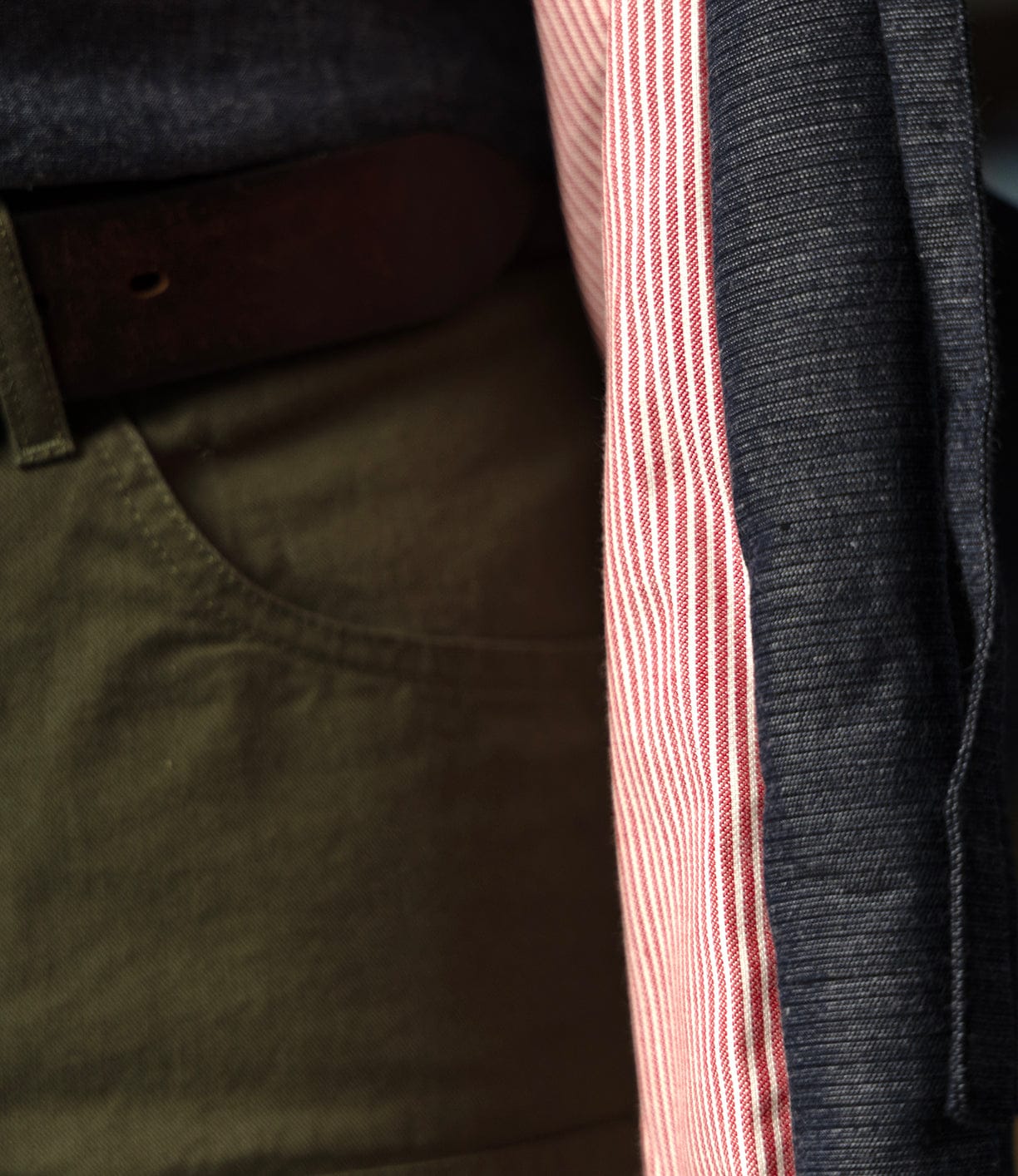 Close-up of a person wearing olive green pants, brown belt, red and white striped shirt, and the Best Made Co. Quilted Hanten Jacket inspired by Japanese workwear; only part of the outfit and pocket are visible.
