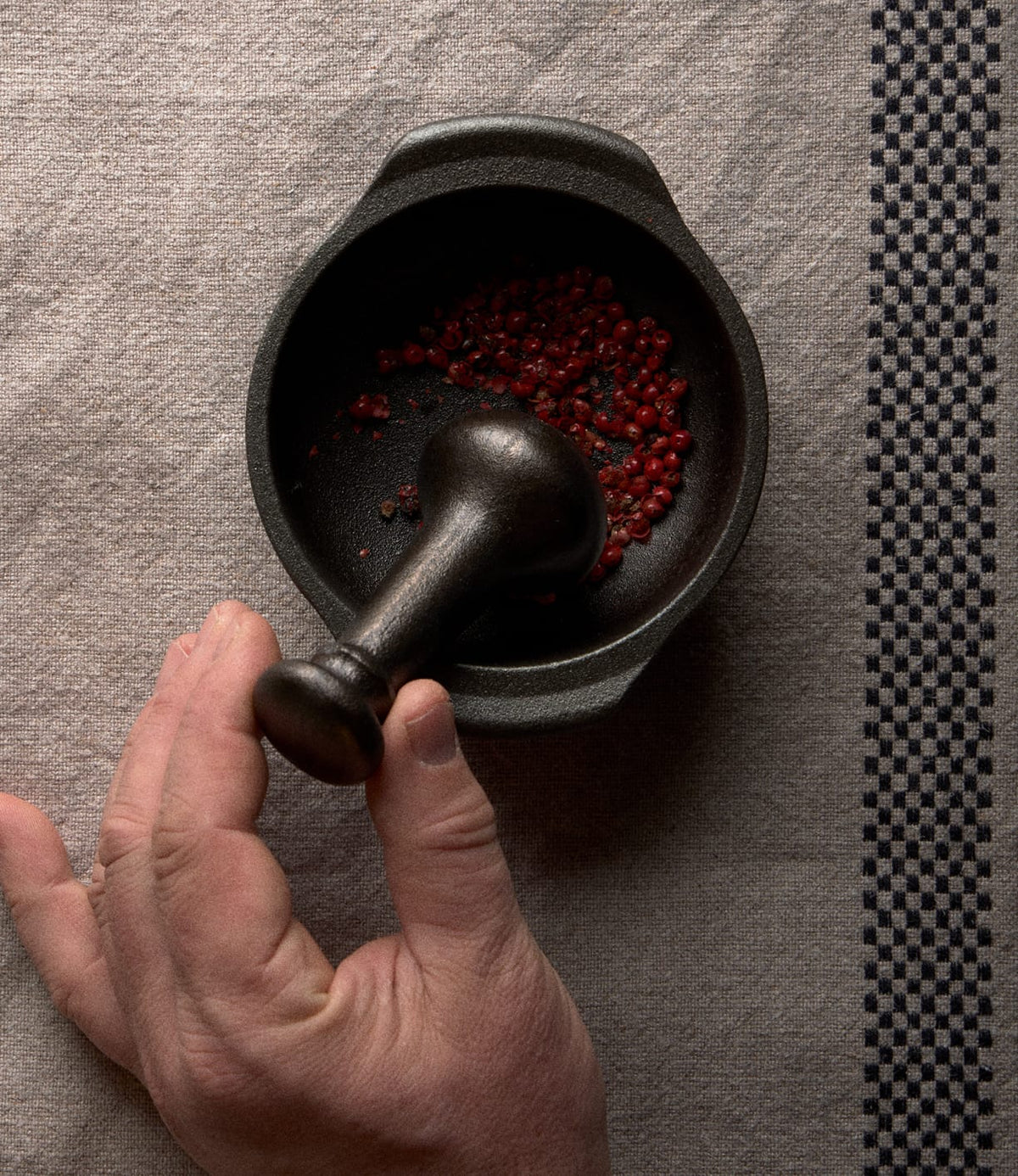 A hand uses a pestle to grind red peppercorns inside a Best Made Co. Cast Iron Mortar and Pestle.