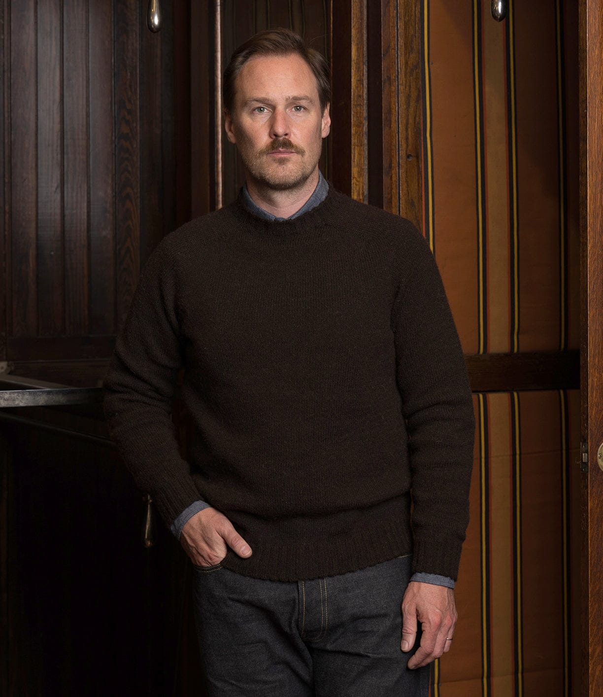 A man with light brown hair and a mustache stands indoors, wearing the Best Made Co. Black Sheep Sweater over a collared shirt and dark jeans. He faces the camera with a neutral expression, framed by dark wood panels and striped wallpaper.