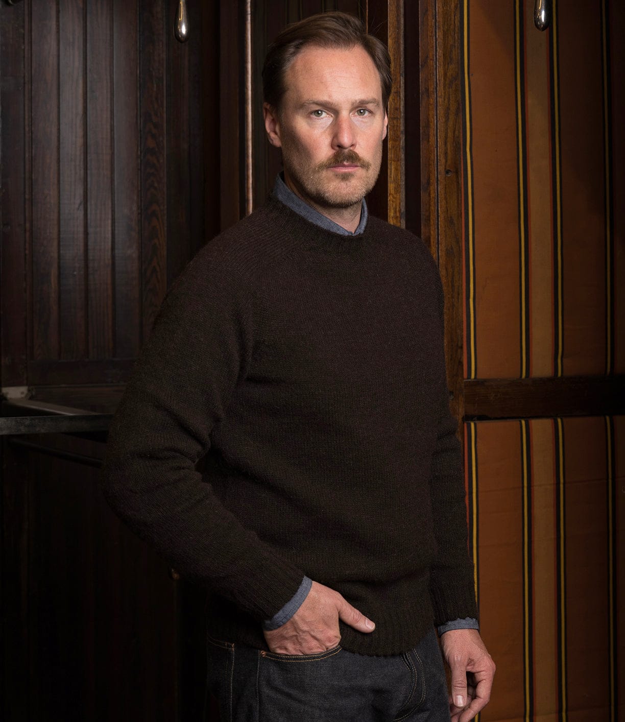 A man with light brown hair and a mustache stands indoors, wearing the Best Made Co. Black Sheep Sweater over a collared shirt and jeans. With one hand in his pocket, he gazes calmly at the camera against dark wood paneling.