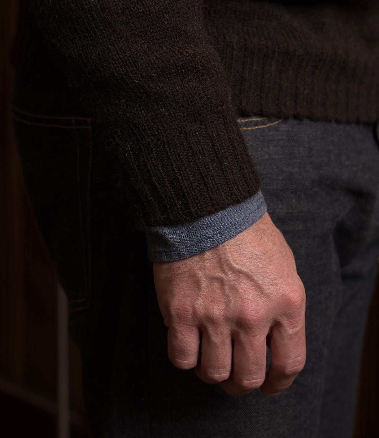 A close-up of a person’s right hand by their leg, wearing dark gray pants and the Best Made Co. Black Sheep Sweater in brown over a blue shirt, highlighting its seamless Shetland wool construction.
