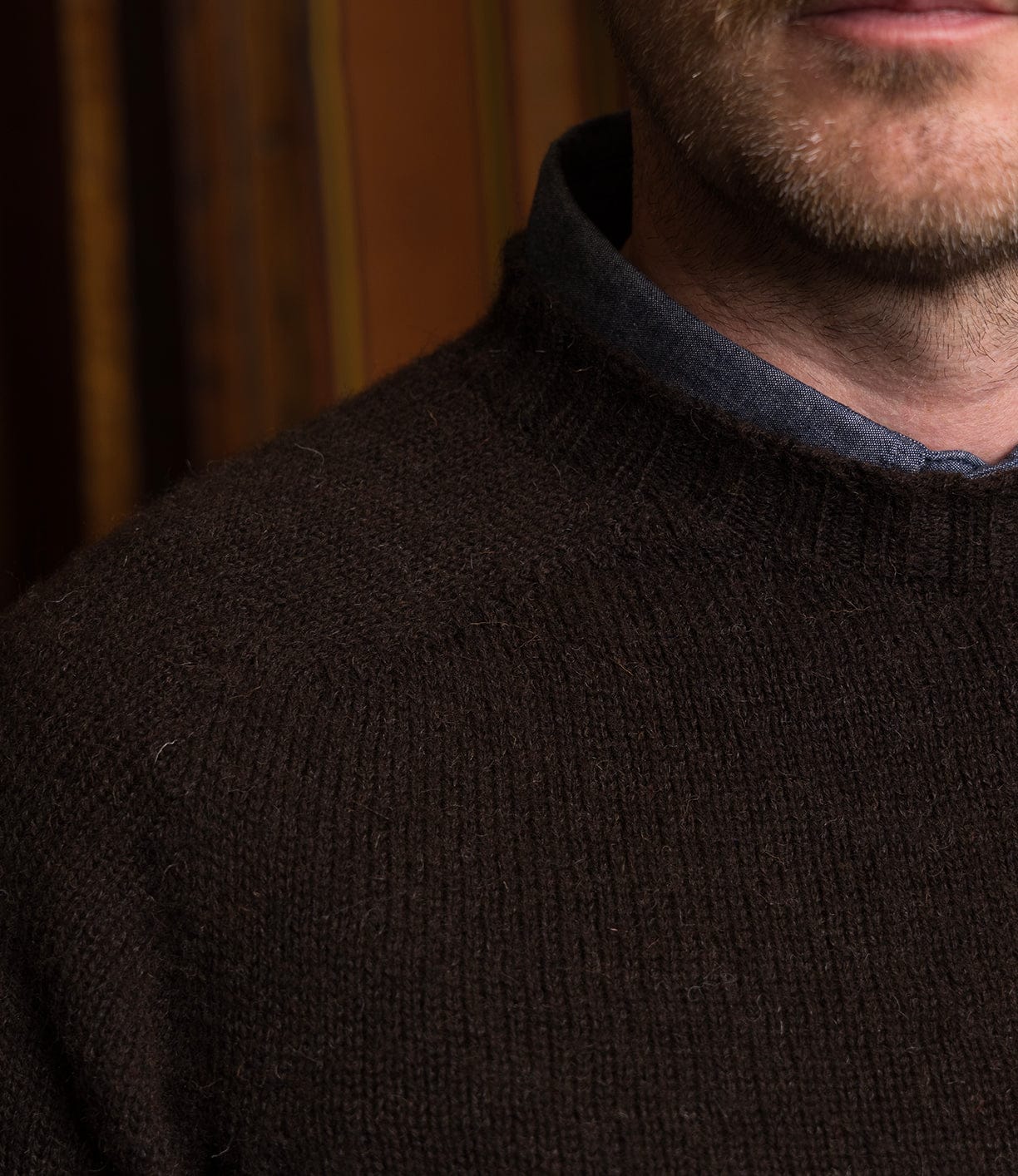 A close-up of a man’s mouth, light stubble, neck, and upper chest in a Best Made Co. Black Sheep Sweater over a blue collared shirt, set against a brown background.