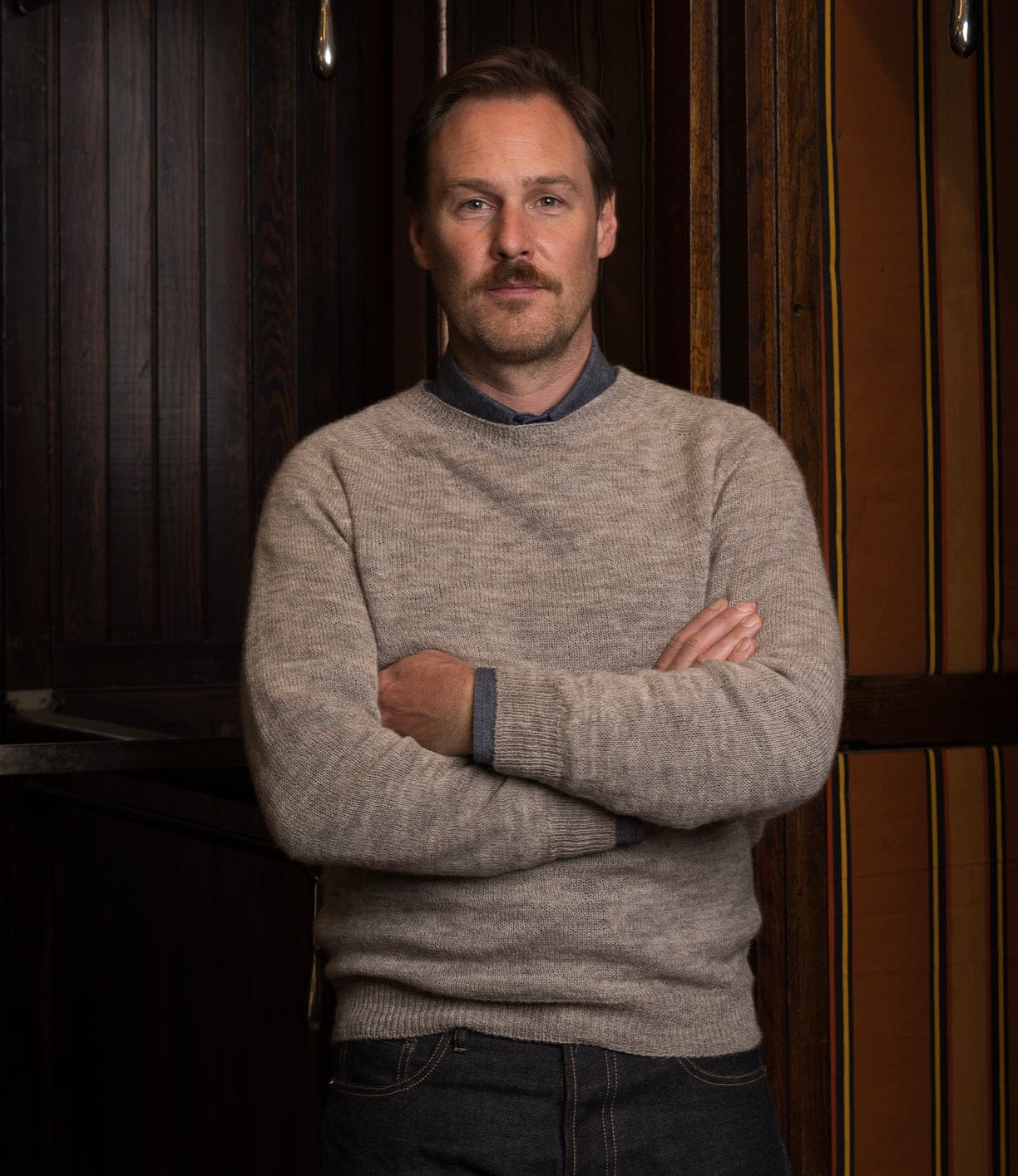 A man with short light brown hair and a mustache stands indoors, arms crossed, wearing the Best Made Co. 5-Gauge Shetland Sweater over a blue collared shirt and dark jeans, in front of wooden paneled walls.