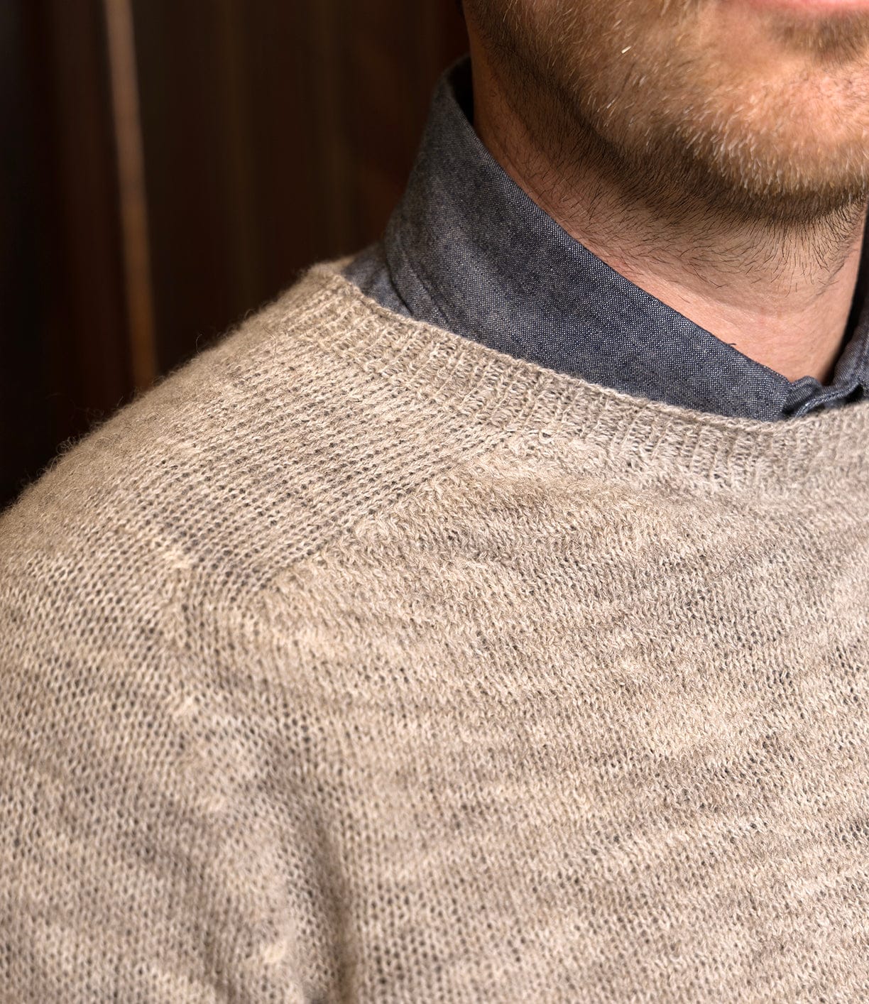 Close-up of a man wearing Best Made Co.’s 5-Gauge Shetland Sweater in light beige over a blue shirt. The blurred, wood-toned background highlights the sweater’s lightweight warmth and classic knit texture.