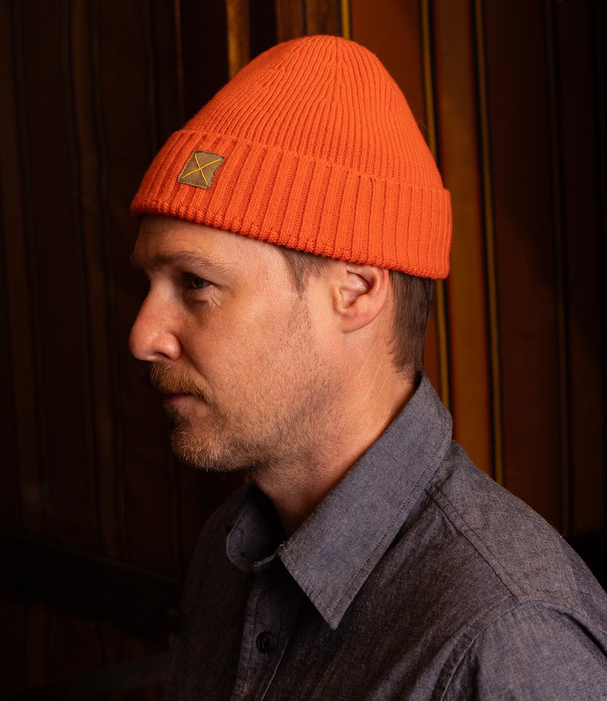 A light-skinned, short-bearded man in profile wears Best Made Co’s orange Cap of Courage and a blue button-up shirt against a dark striped background.