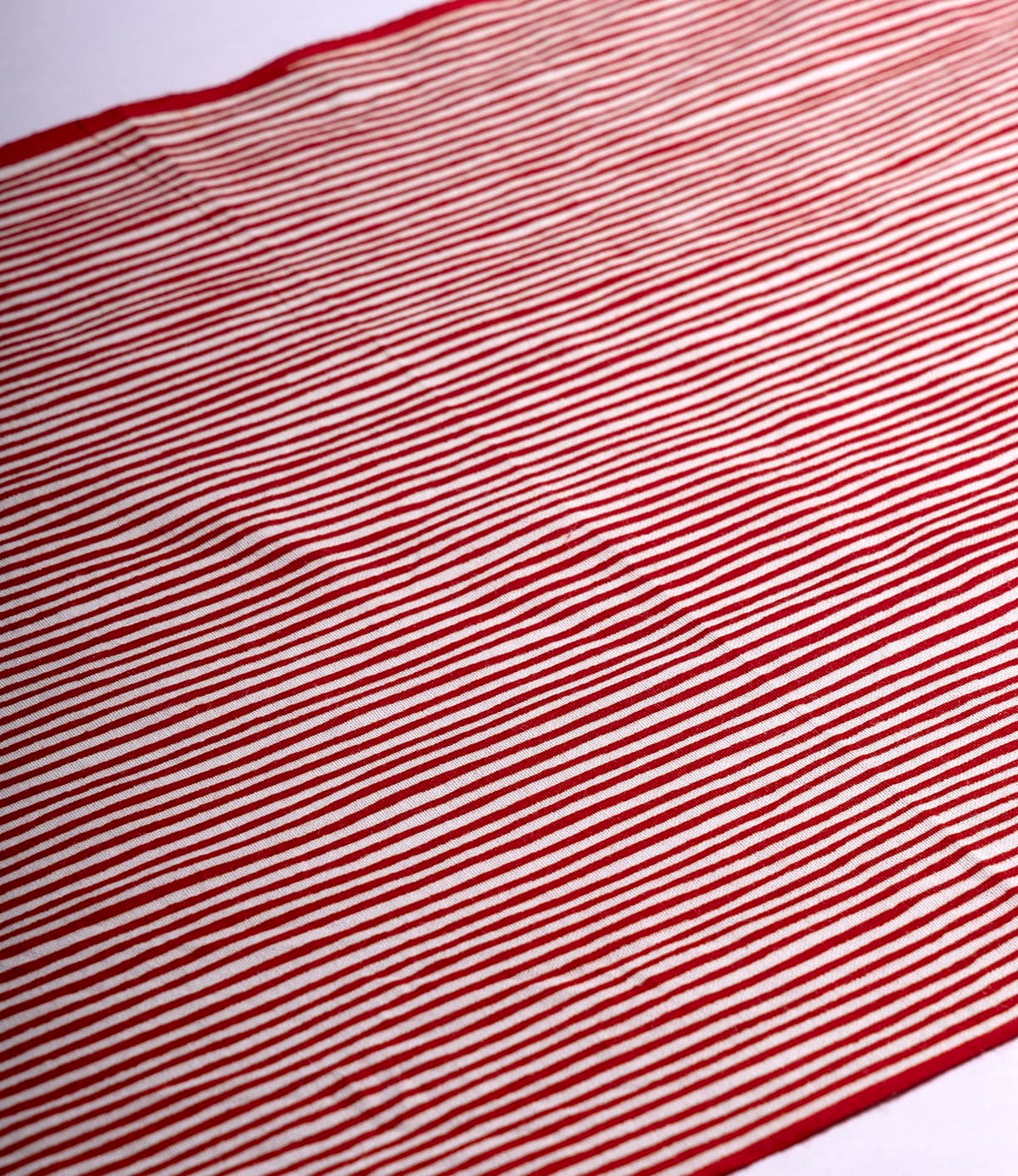 The Yoroke-Jima Tenugui by Best Made Co. features red and white horizontal stripes on hand-dyed Japanese fabric, laid flat with gentle wrinkles and shadows for a wabi-sabi feel. Evenly spaced stripes span the entire cloth.