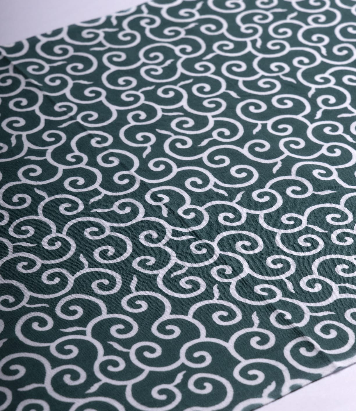 The Karakusa Tenugui by Best Made Co. is a hand-dyed Japanese cloth featuring a repeating white swirl on dark green in a diagonal pattern, inspired by wabi-sabi aesthetics.