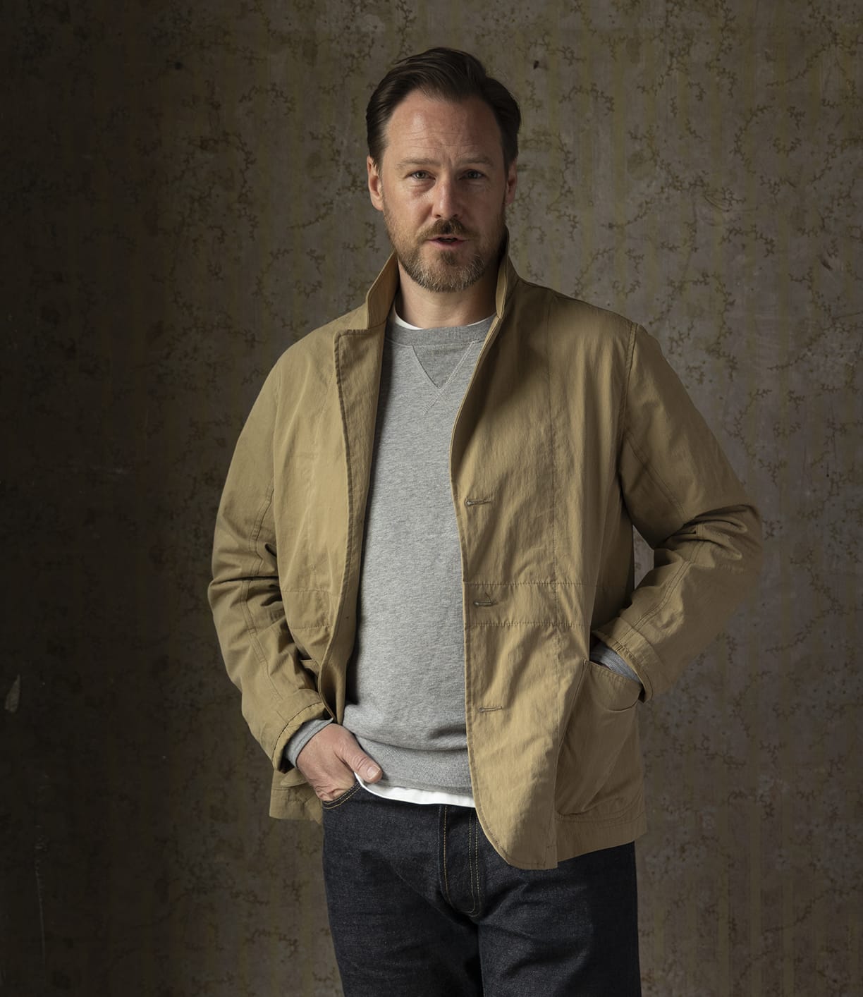 A man with short brown hair and a beard stands against a textured wall, wearing the Best Made Co. Possibilities Jacket Tan over a gray sweatshirt and dark jeans, with one hand in his jacket pocket.