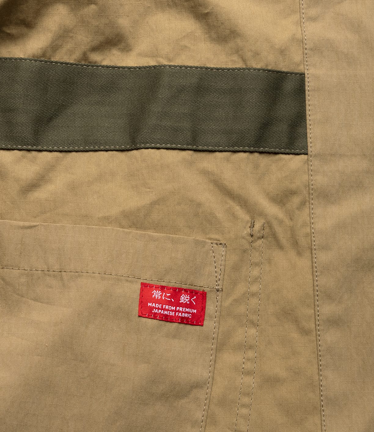 A close-up of the Possibilities Jacket Tan by Best Made Co.—khaki fabric with a green stripe, pocket, and red label in Japanese reading "Made from Premium Japanese Fabric," highlighting its durable, lightweight craftsmanship.
