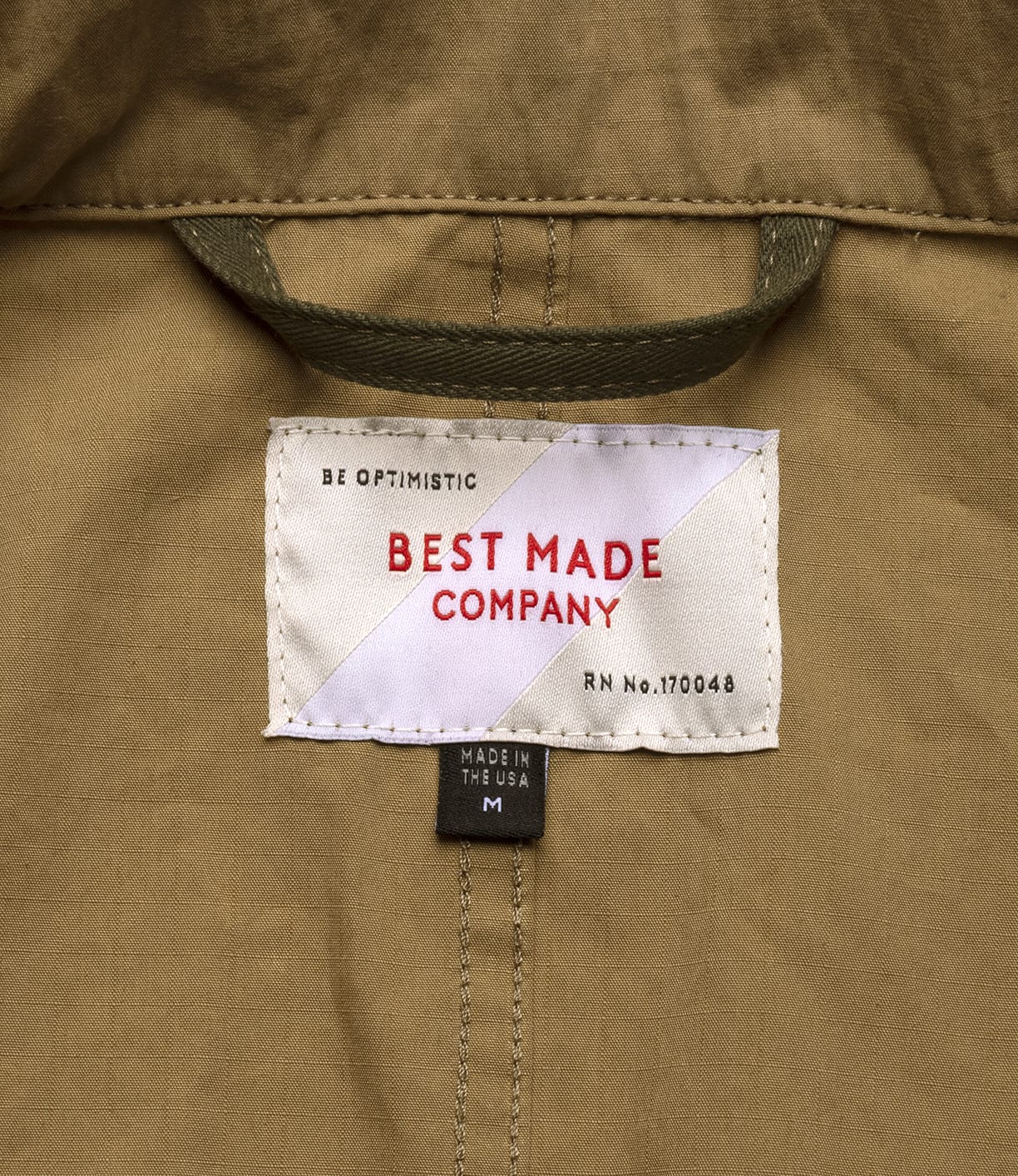 A close-up of a label on tan fabric from the Best Made Co. Possibilities Jacket Tan, reading BEST MADE COMPANY in red, BE OPTIMISTIC above, and smaller tags below that state MADE IN THE USA and M.